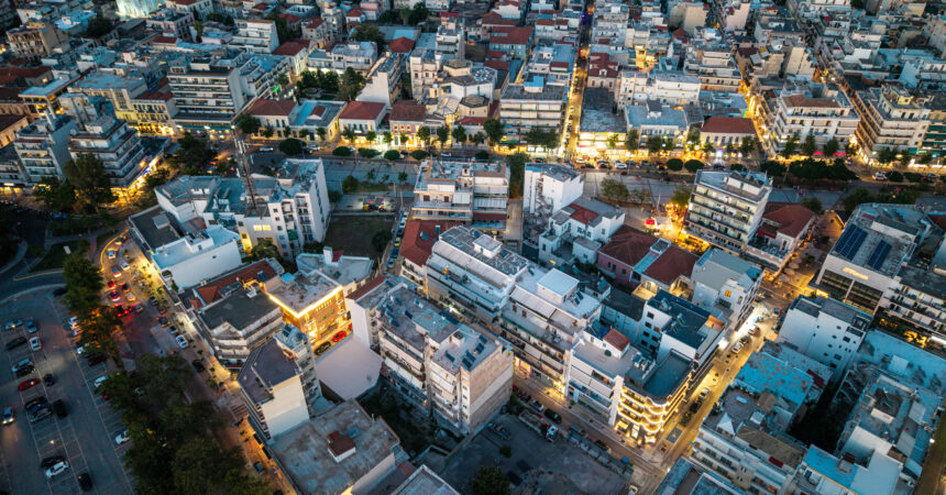 Aerial view of a dense city block at dusk, with many multi-story buildings, narrow streets, and illuminated storefronts.