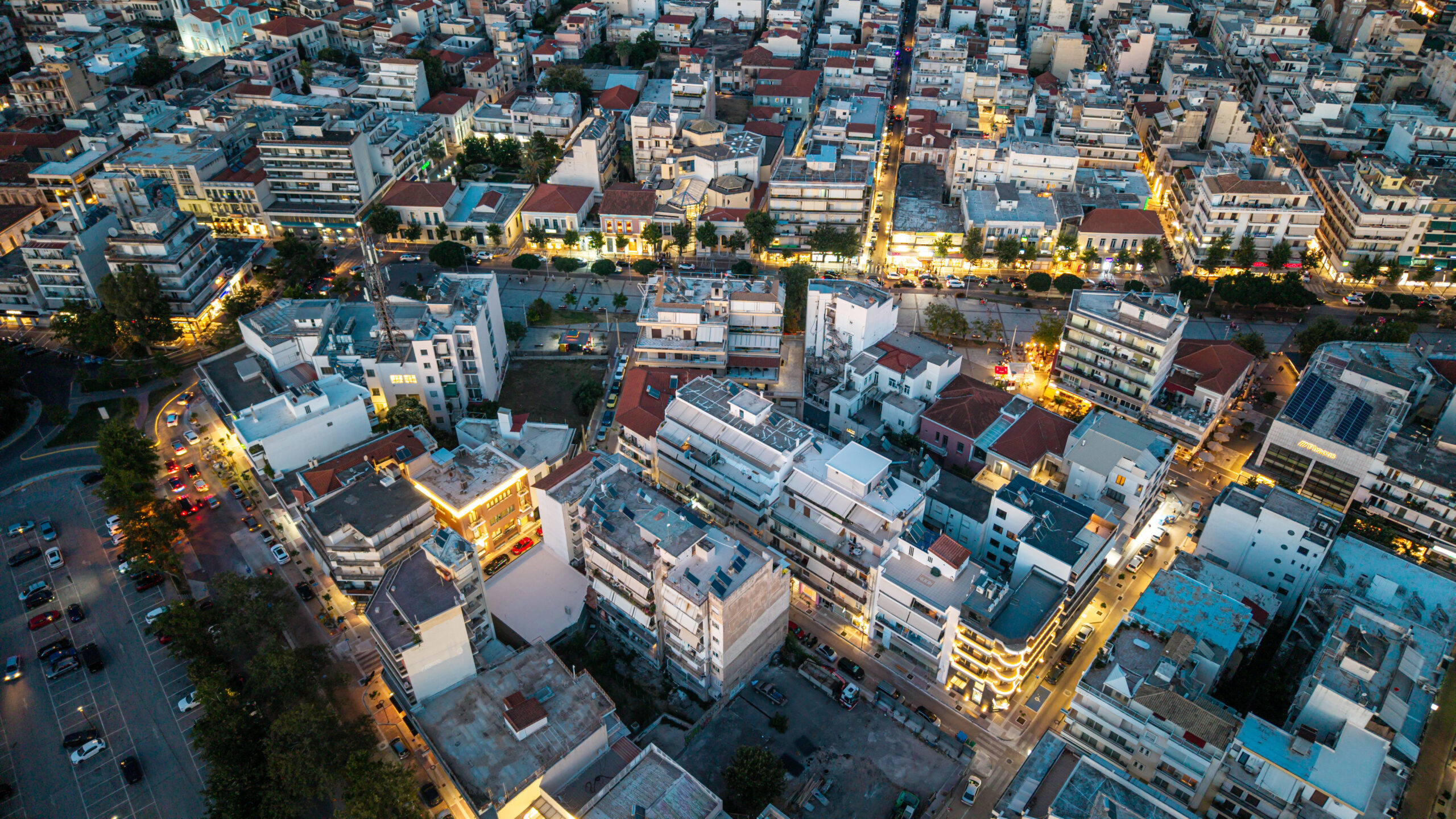 Aerial view of a dense city block at dusk, with many multi-story buildings, narrow streets, and illuminated storefronts.